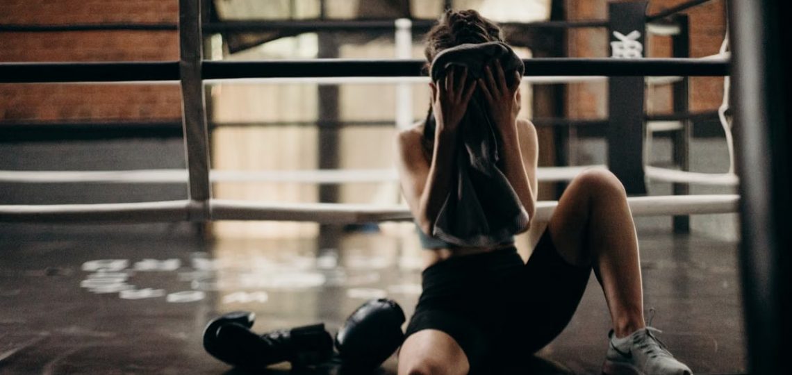 A female boxer sitting in a gym ring, resting and wiping sweat with a towel after a workout.